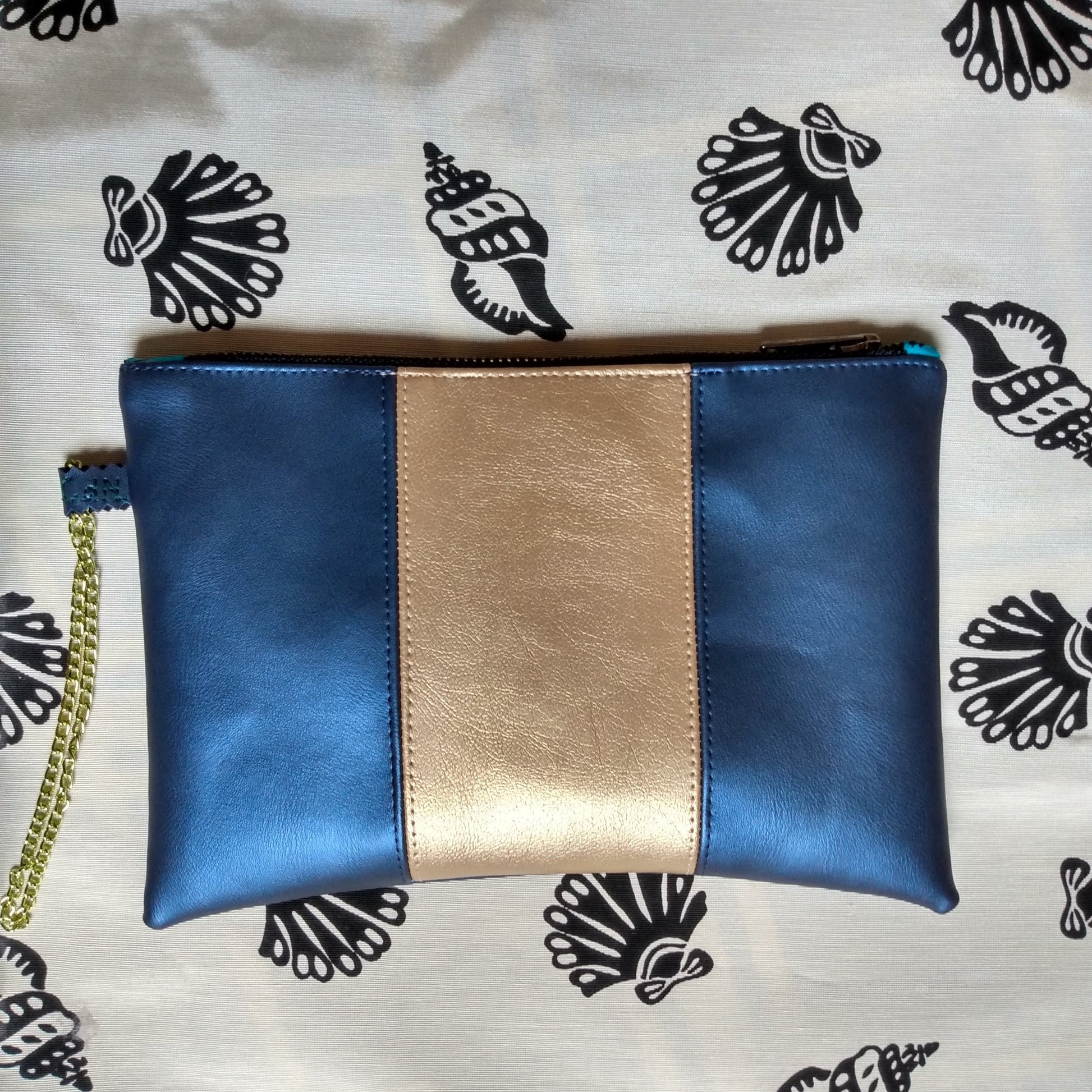Vue avant pochette Soglad en simili cuir. 3 bandes verticales : bleu métallisé, doré et bleu métallisé. Une dragonne en chainette permet le port de la pochette au poignet. Une fermeture éclair ferme la pochette.