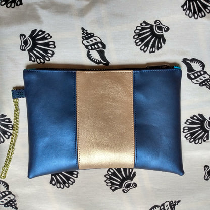 Vue avant pochette Soglad en simili cuir. 3 bandes verticales : bleu métallisé, doré et bleu métallisé. Une dragonne en chainette permet le port de la pochette au poignet. Une fermeture éclair ferme la pochette.