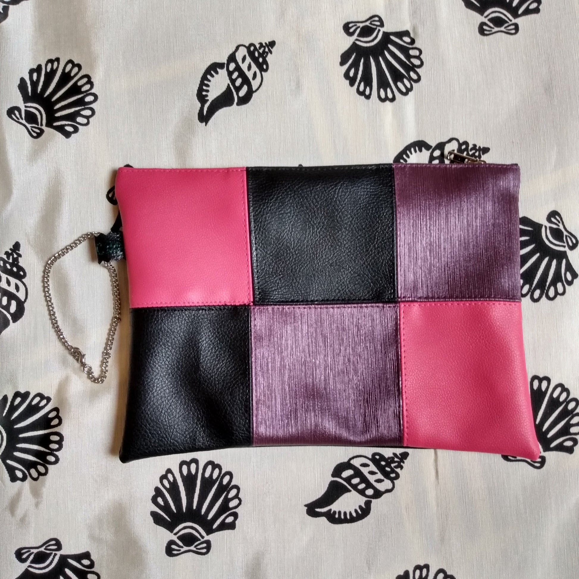 Pochette de soirée en simili cuir, vue avant. Damier 6 carreaux de couleur rose, noir et violet. Une dragonne sur un côté permet le port de la pochette à la main. Se ferme grâce à une fermeture éclair.