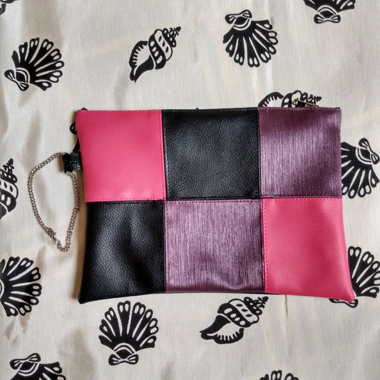 Pochette de soirée en simili cuir, vue avant. Damier 6 carreaux de couleur rose, noir et violet. Une dragonne sur un côté permet le port de la pochette à la main. Se ferme grâce à une fermeture éclair.
