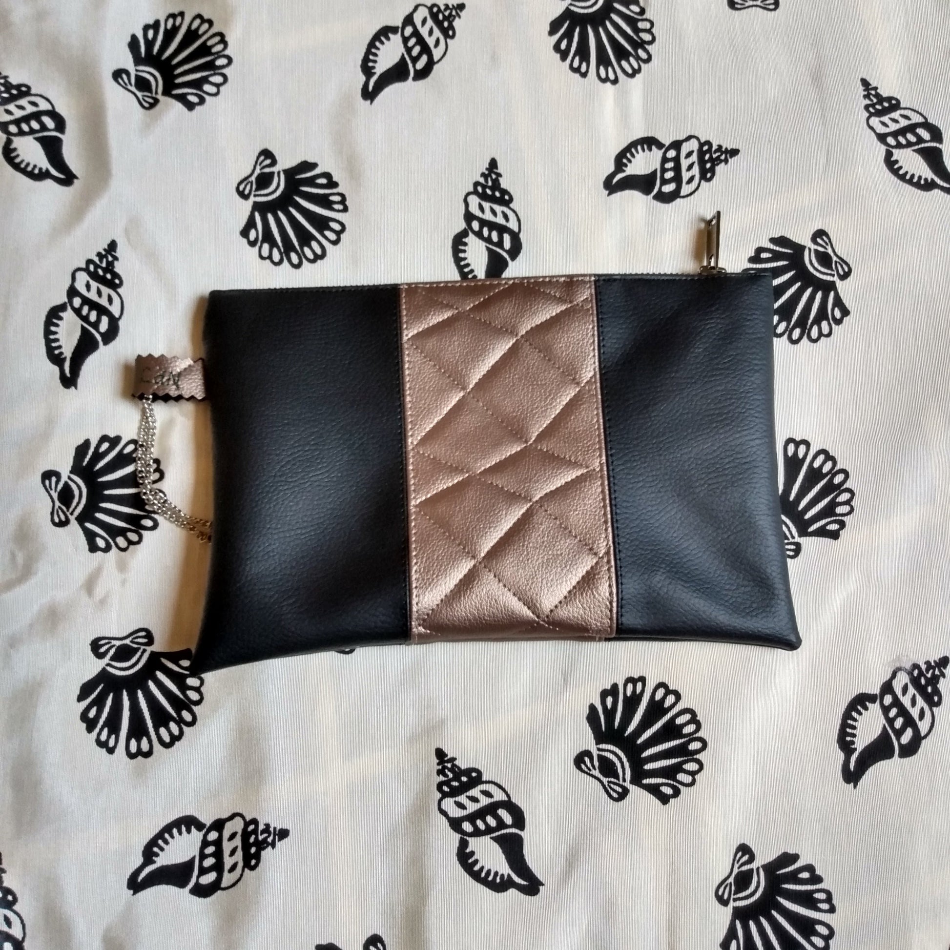 Vue avant pochette Soglad. 3 bandes simili cuir verticales : noir, doré effet matelassé et noir. Une dragonne en chainette permet le port de la pochette au poignet. Une fermeture éclair ferme la pochette.