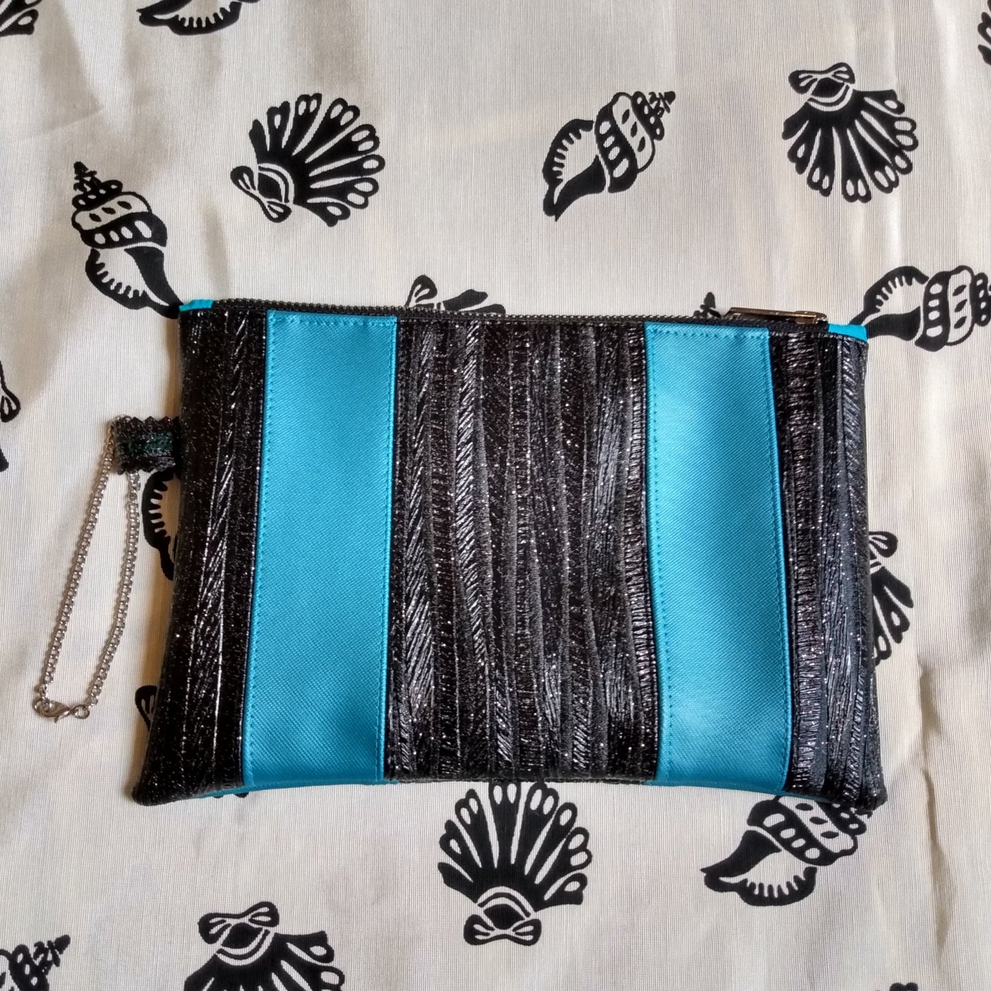 Vue avant pochette Soglad. 5 bandes verticales qui alternent entre noir pailleté et bleu canard. Une dragonne en chainette permet le port de la pochette au poignet. Une fermeture éclair ferme la pochette.