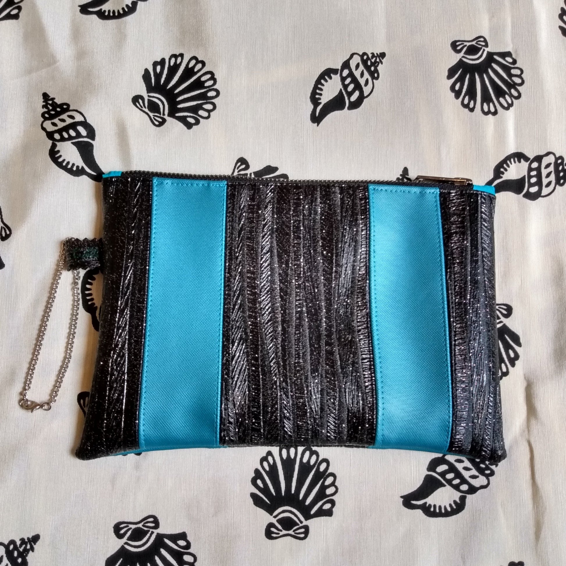 Vue avant pochette Soglad. 5 bandes verticales qui alternent entre noir pailleté et bleu canard. Une dragonne en chainette permet le port de la pochette au poignet. Une fermeture éclair ferme la pochette.