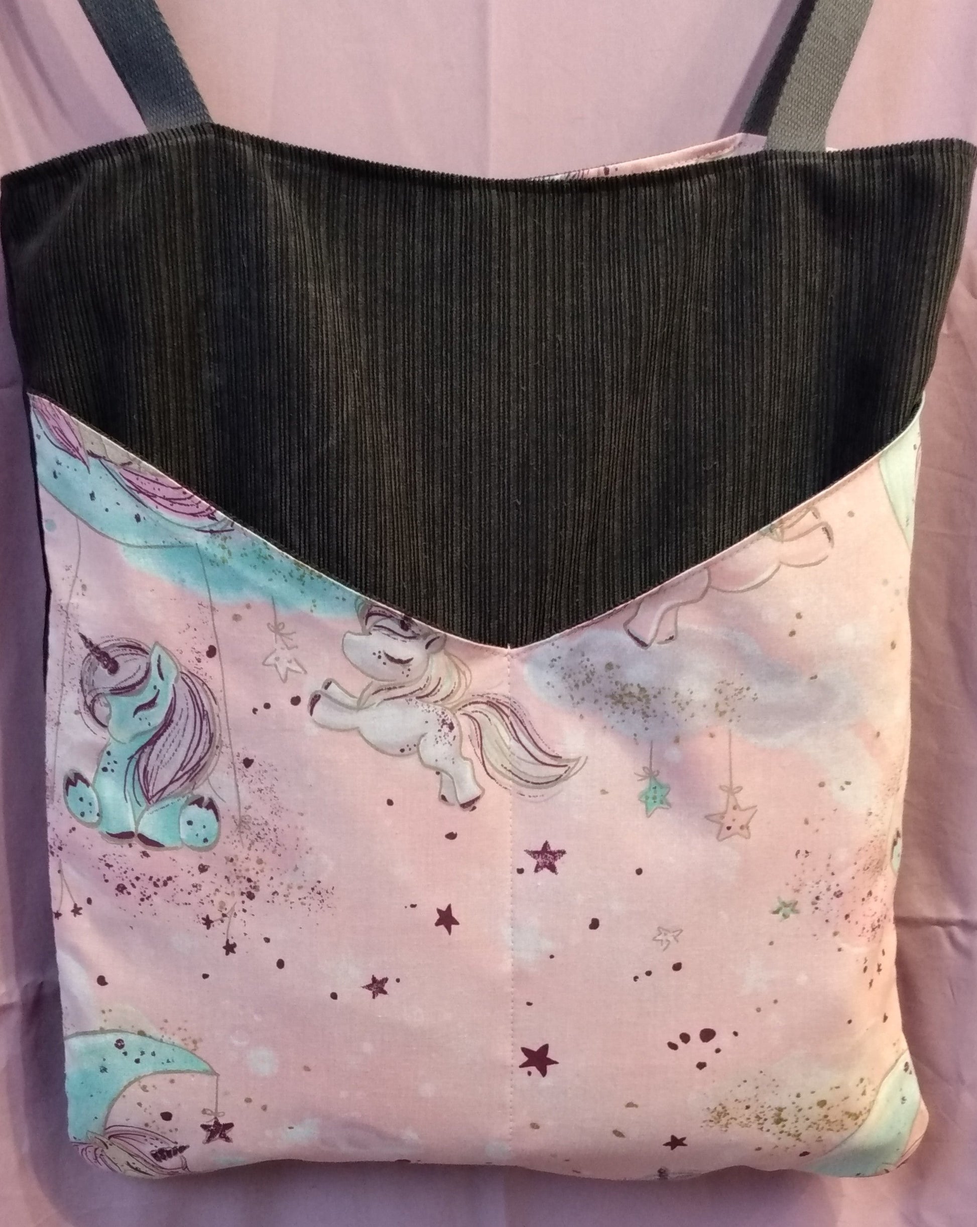 Avant du sac Balad'In rose et gris, tissu principal velours gris anthracite avec deux poches plaquées, qui partent du bas jusqu'aux 3/4 du sac environ. Poches motif licornes et étoiles couleurs pâles. Les anses sont faites pour porter le sac à l'épaule et sont en coton gris.