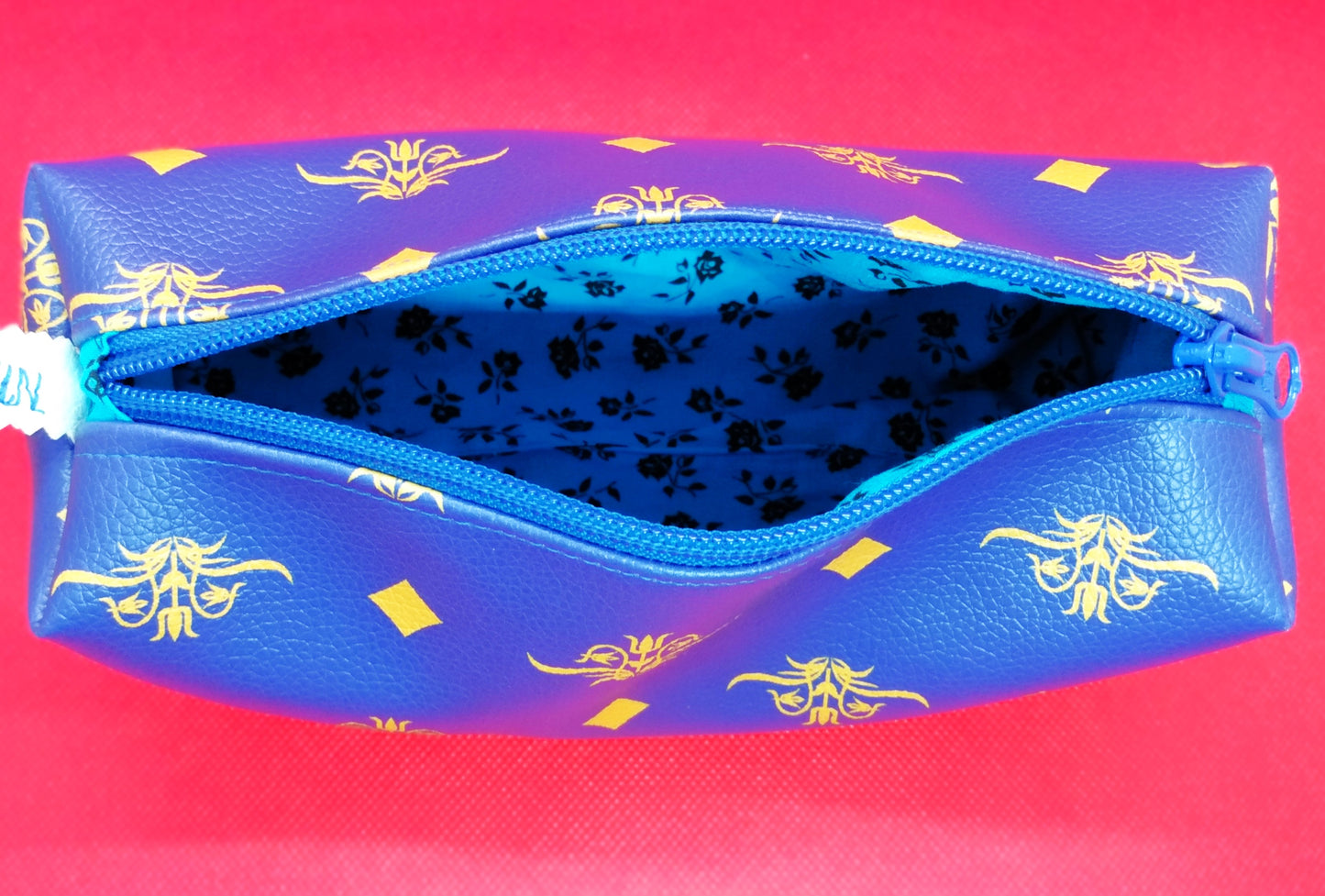 Trousse en simili cuir bleu roi avec fleurs et losanges jaunes. La trousse est ouverte. On voit la doublure intérieure avec des roses noires sur fond bleu clair.