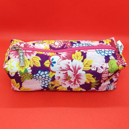 Trousse taille 1 tissu à grosses fleurs roses, jaunes, bleues ou vertes, sur fond violet foncé. Se ferme par une fermeture éclair rose. L'étiquette "CdN" de la marque est présente sur un côté de la trousse.