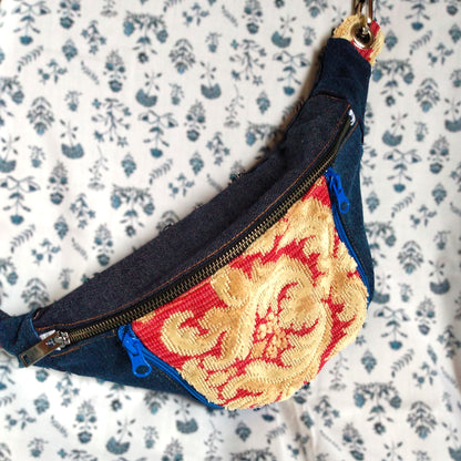 Avant du sac banane Mango. Tissu principal extérieur Jean bleu marine. Une grande fermeture métal pour ouvrir la poche principale. Une poche avant est présente au centre de la banane. La poche est en tissu jacquard rouge avec un décor jaune paille. Cette poche s'ouvre via deux fermetures éclair bleu roi. On aperçoit que la sangle s'accroche par des mousquetons métal reliés à des œillets.