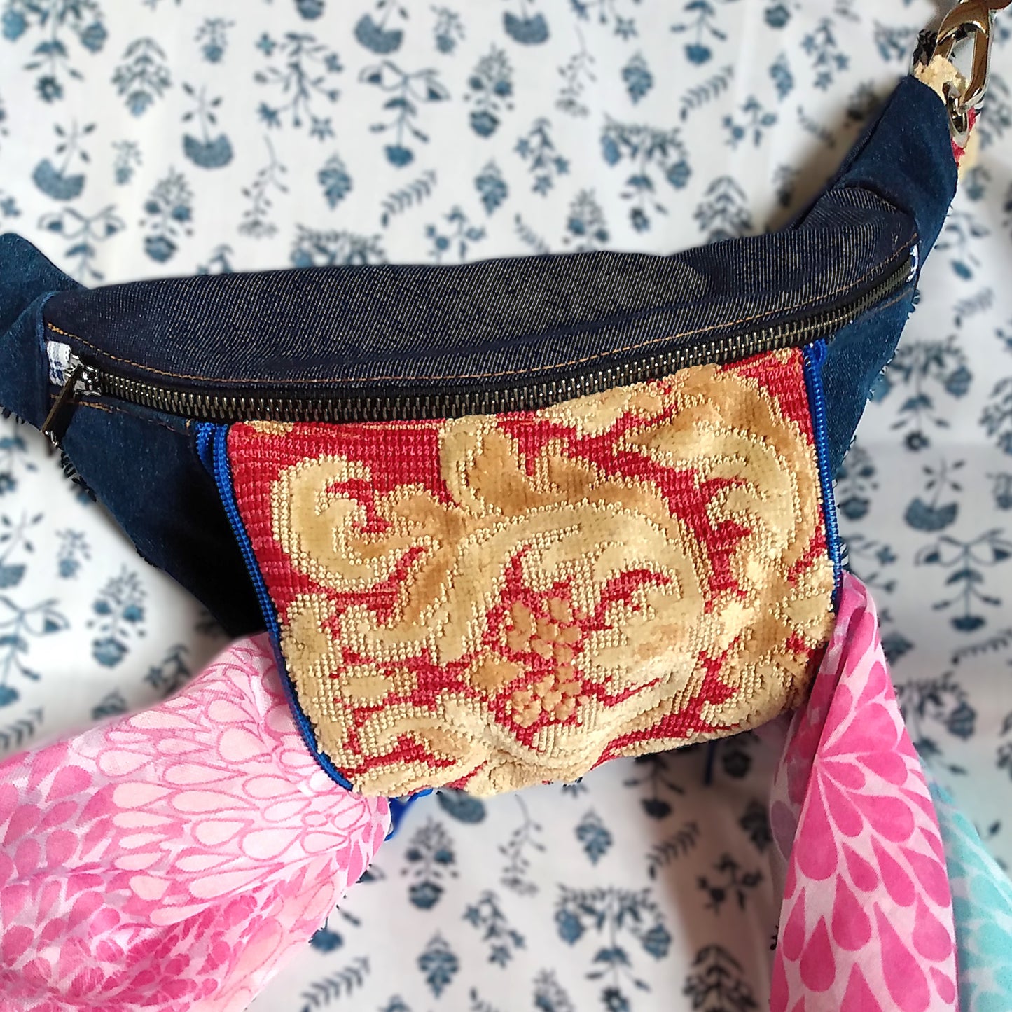 Vue avant du sac Mango. Tissu principal extérieur Jean bleu marine. Une grande fermeture métal pour ouvrir la poche principale. Une poche avant est présente au centre de la banane. La poche est en tissu jacquard rouge avec un décor jaune paille. Cette poche s'ouvre via deux fermetures éclair bleu roi. Les deux fermetures sont ouvertes et une écharpe rose est placée à l'intérieur en dépassant des deux côtés. Cela montre que le poche peut s'ouvrir à gauche et/ou à droite.