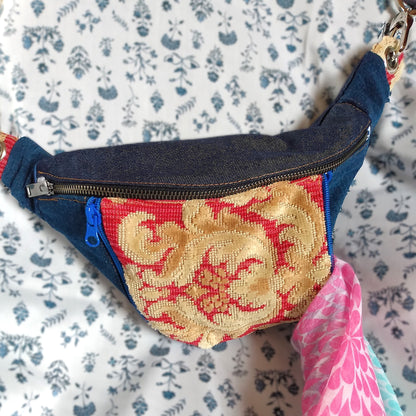 Avant du sac banane Mango. Tissu principal extérieur Jean bleu marine. Une grande fermeture métal pour ouvrir la poche principale. Une poche avant est présente au centre de la banane. La poche est en tissu jacquard rouge avec un décor jaune paille. Cette poche s'ouvre via deux fermetures éclair bleu roi. La poche est ouverte d'un côté et laisse entrevoir une écharpe rose. On aperçoit que la sangle s'accroche par des mousquetons métal reliés à des œillets.