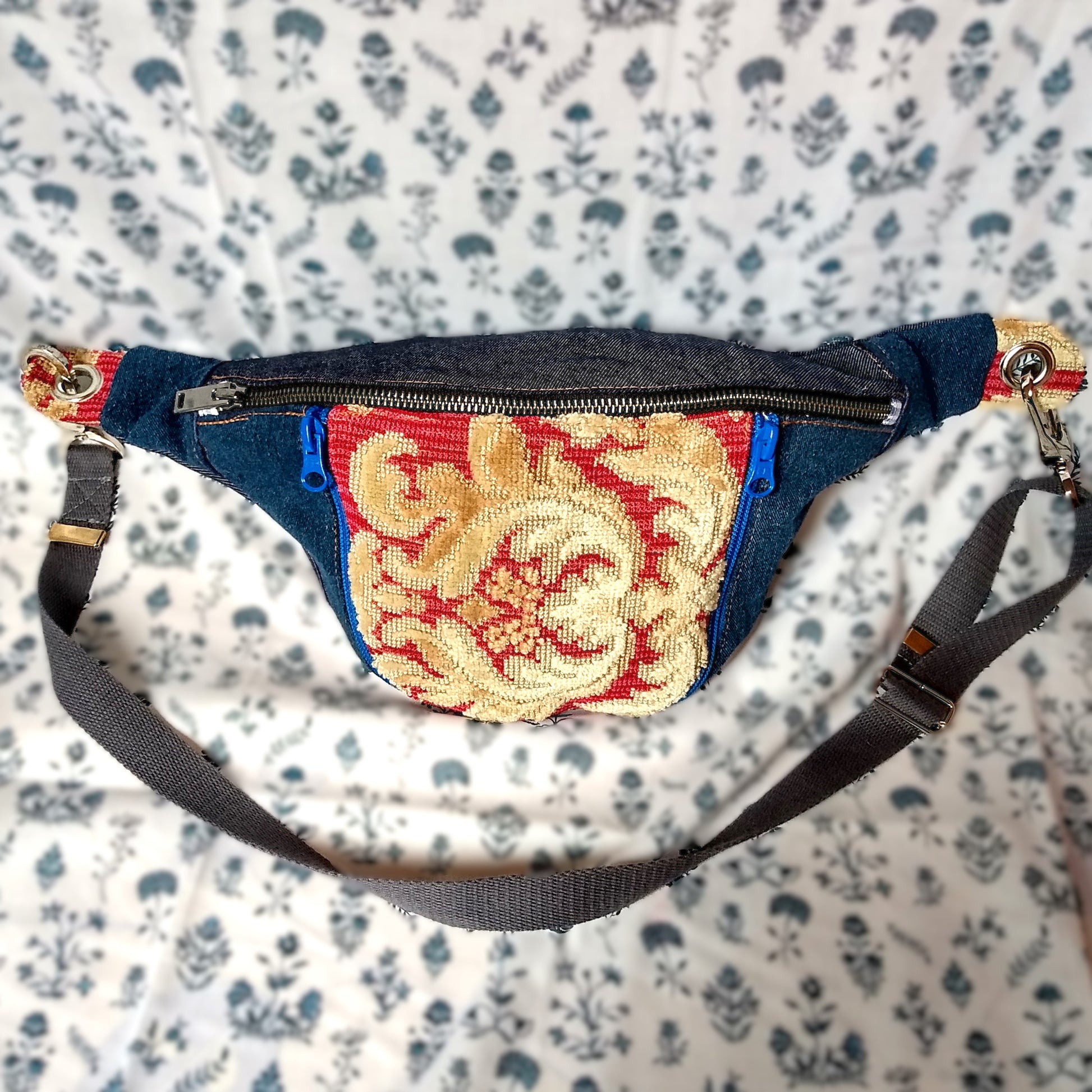 Avant du sac banane Mango. Tissu principal extérieur Jean bleu marine. Une grande fermeture métal pour ouvrir la poche principale. Une poche avant est présente au centre de la banane. La poche est en tissu jacquard rouge avec un décor jaune paille en velours. Cette poche s'ouvre via deux fermetures éclair bleu roi, une fermeture de chaque côté de la poche. La sangle réglable est attachée via des mousquetons, elle est donc amovible. La sangle est en coton gris.