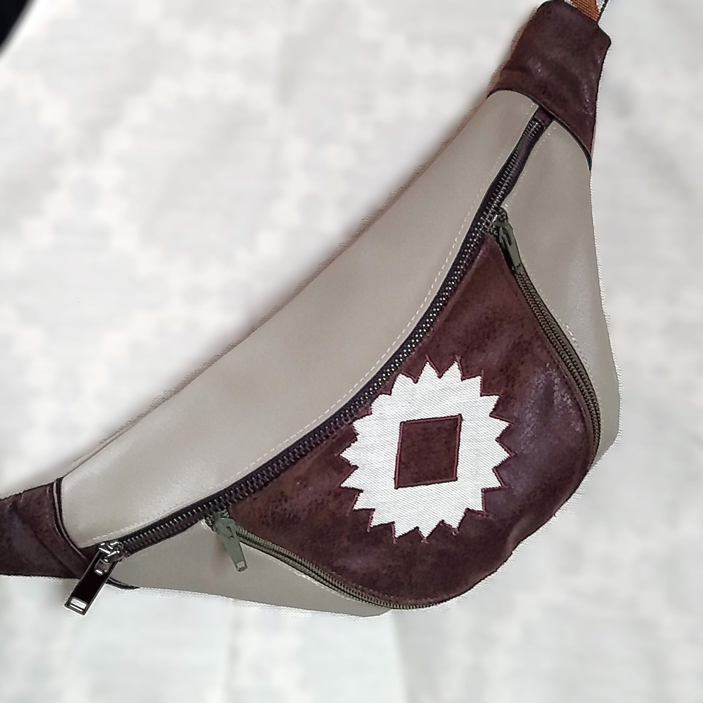 Avant du sac banane Mango. Tissu principal extérieur simili cuir beige. Une grande fermeture métal pour ouvrir la poche principale. Une poche avant est présente au centre de la banane. La poche est en simili marron foncé aspect daim avec au centre un appliqué en forme de soleil beige. Cette poche s'ouvre via deux fermetures vert kaki.