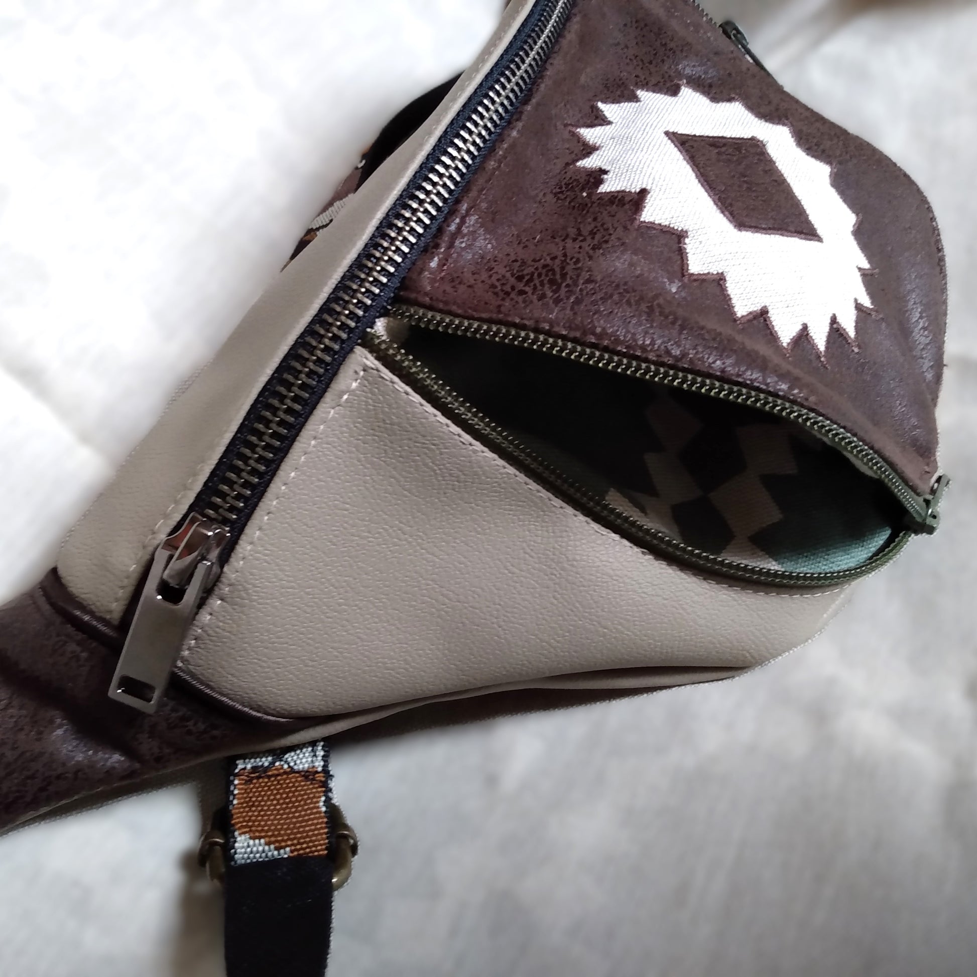 Vue de côté. Tissu principal extérieur simili cuir beige. Une grande fermeture métal pour ouvrir la poche principale. La poche avant est en simili marron foncé aspect daim avec au centre un appliqué en forme de soleil beige. Cette poche est ouverte via une fermeture vert kaki. Cela laisse entrevoir la doublure de la poche verdé et beige.