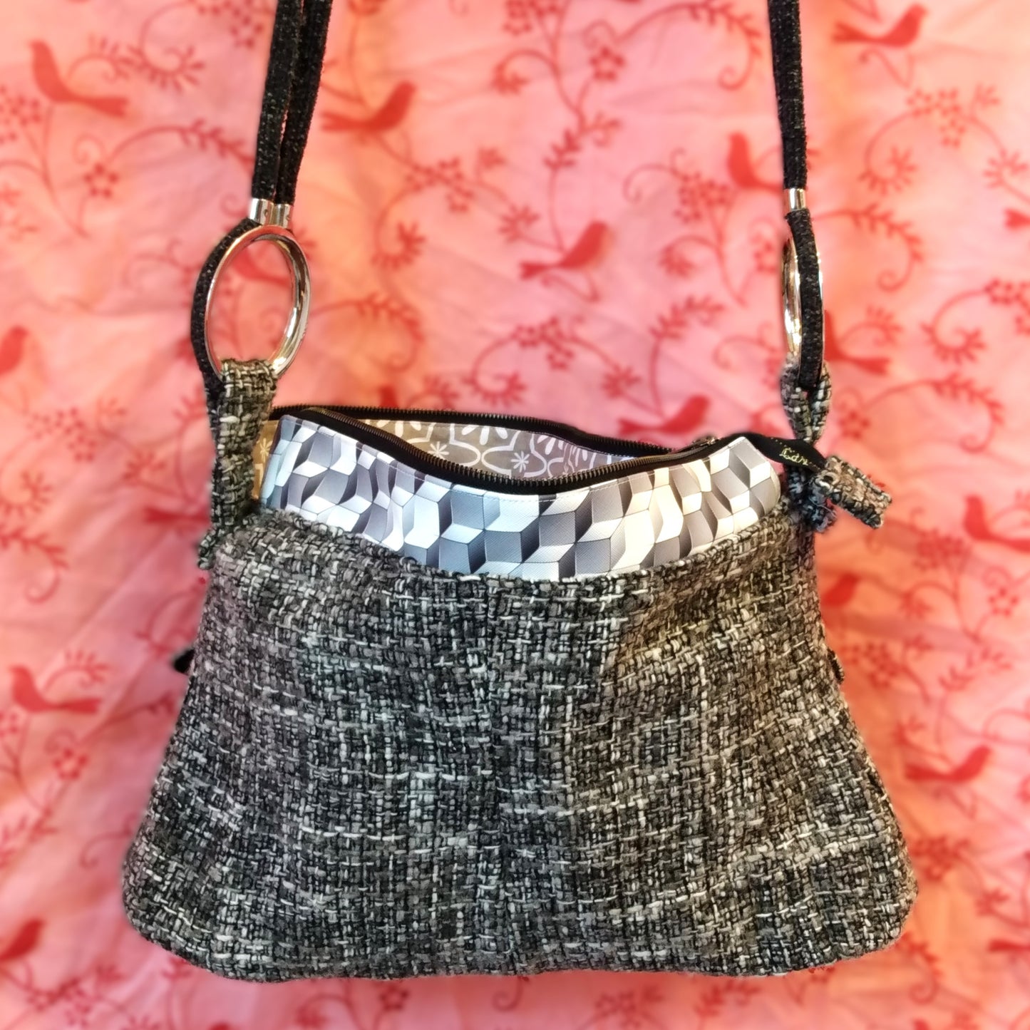 Arrière du sac Laina. Avant du sac Laina. Le sac est majoritairement en laine grise noire et blanche. Le haut du sac possède une bande horizontale en similicuir motif cubes gris et blancs. Le sac se ferme via une grande fermeture éclair métal. L'anse du sac est accrochée sur les côtés du sac via de gros anneau en métal. Elle est en velours gris anthracite.