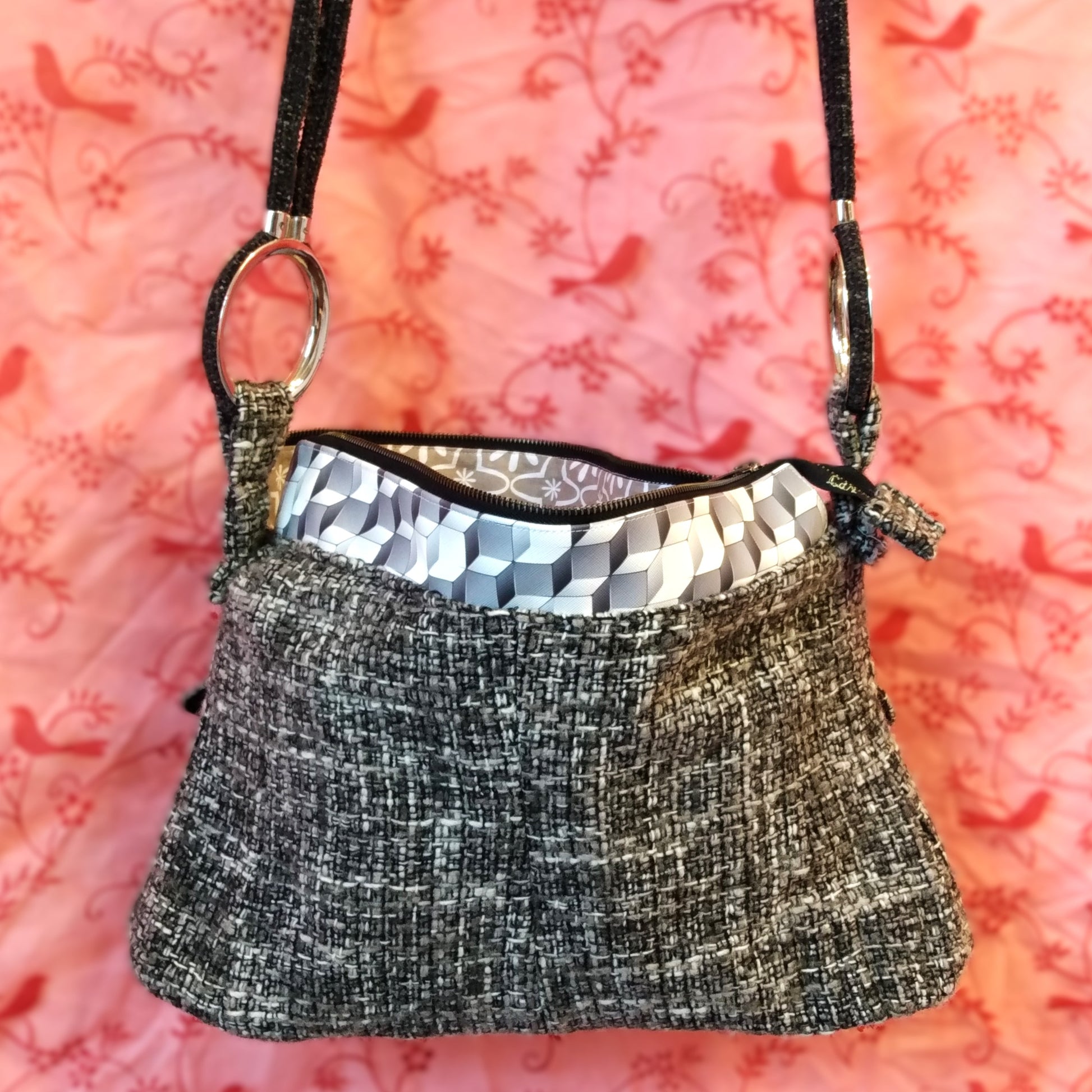 Arrière du sac Laina. Avant du sac Laina. Le sac est majoritairement en laine grise noire et blanche. Le haut du sac possède une bande horizontale en similicuir motif cubes gris et blancs. Le sac se ferme via une grande fermeture éclair métal. L'anse du sac est accrochée sur les côtés du sac via de gros anneau en métal. Elle est en velours gris anthracite.