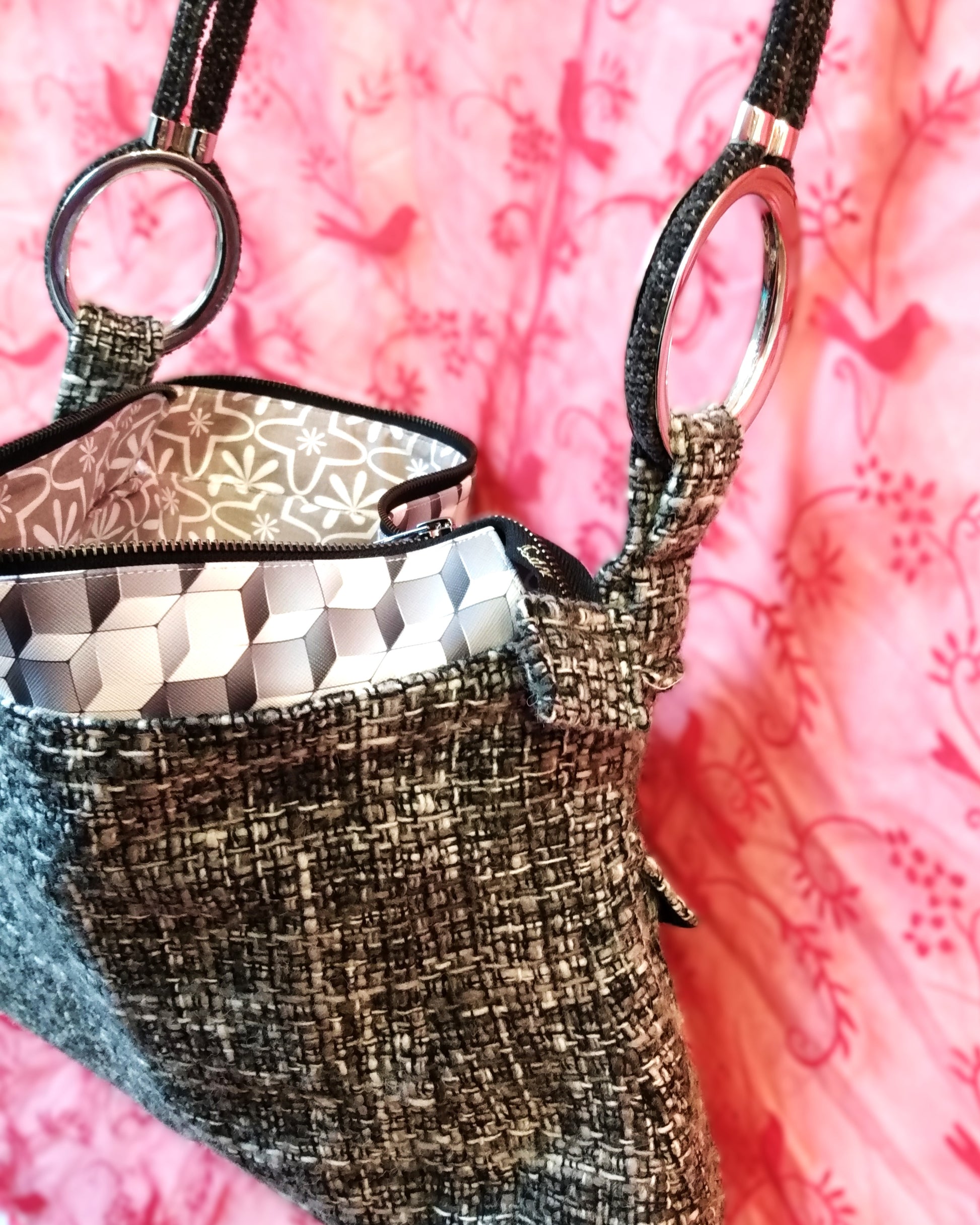 Vue de côté du sac Laina. Avant du sac Laina. Le sac est majoritairement en laine grise noire et blanche. Le haut du sac possède une bande horizontale en similicuir motif cubes gris et blancs. Le sac s'ouvre via une grande fermeture éclair métal. L'anse du sac est accrochée sur les côtés du sac via de gros anneaux en métal. Elle est en velours gris anthracite. La doublure intérieure du sac est grise à motifs floraux blancs.