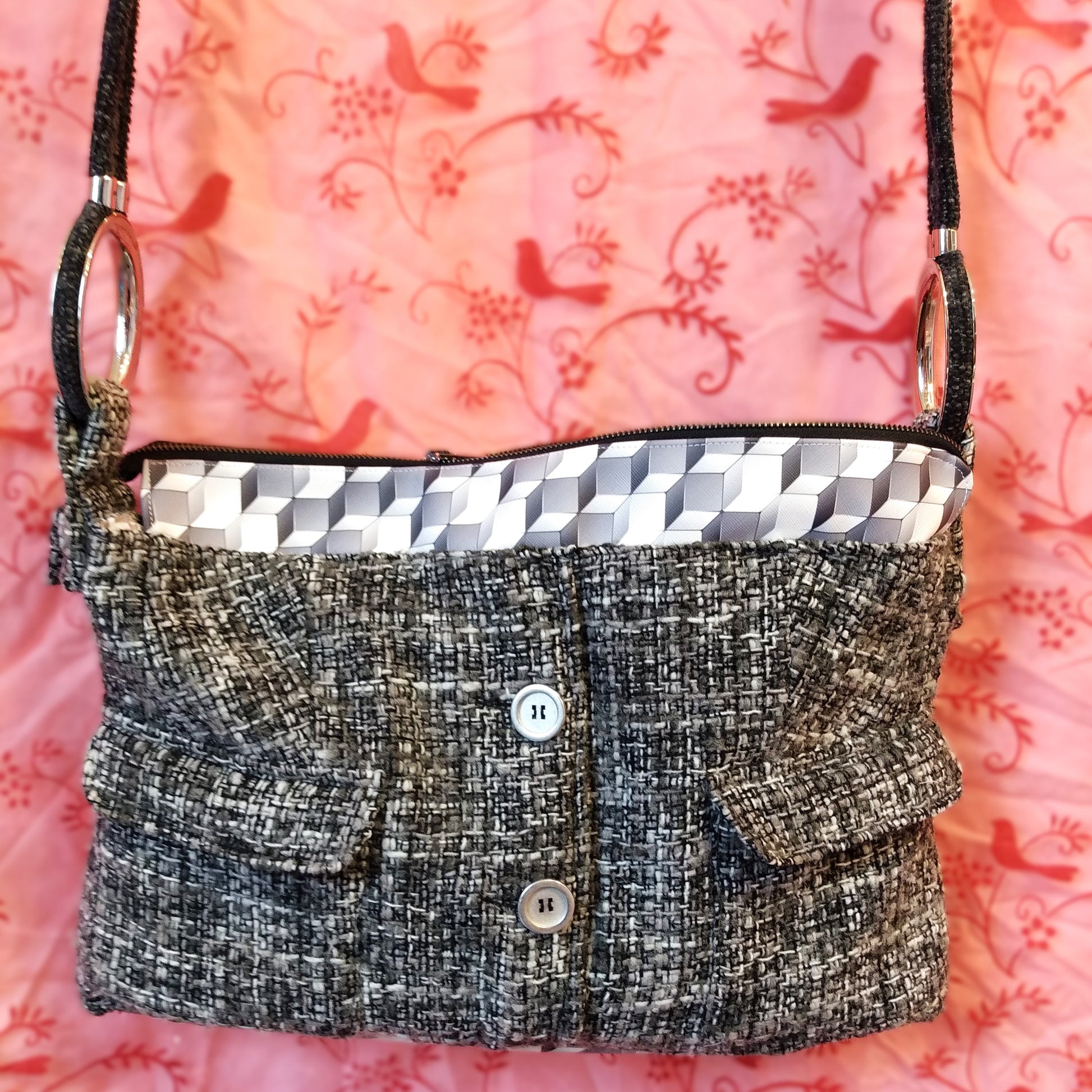 Avant du sac Laina. Le sac est majoritairement en laine grise noire et blanche. On voit deux gros boutons métallisés au centre et deux poches à rabat une de chaque côtés. Le haut du sac possède une bande horizontale en similicuir motif cubes gris et blancs. Le sac se ferme via une grande fermeture éclair métal. L'anse du sac est accrochée sur les côtés du sac via de gros anneaux en métal. Elle est en velours gris anthracite.