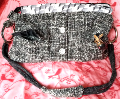 Avant du sac Laina. Le sac est majoritairement en laine grise noire et blanche. On voit deux gros boutons métallisés au centre et deux poches à rabat une de chaque côtés. Le haut du sac possède une bande horizontale en similicuir motif cubes gris et blancs. Le sac se ferme via une grande fermeture éclair métal.L'anse du sac est accrochée sur les côtés du sac via de gros anneaux en métal. Elle est en velours gris anthracite.