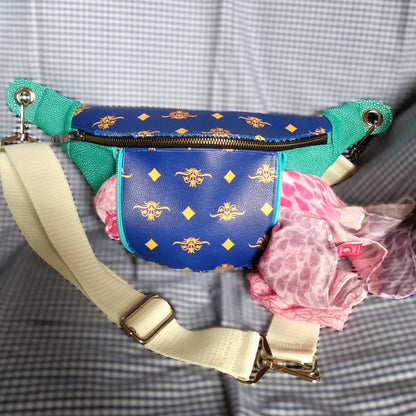 Vue avant du sac banane Mango version bleue. L'extérieur est bicolore en simili cuir émeraude à grains et bleu roi à motifs fleurs et losanges jaunes. Une grande poche principale se ferme via un zip métal. Une poche avant est ouverte par un zip bleu clair de chaque côté. On voit dépasser une écharpe rose des deux côtés de la poche. La sangle est beige et réglable. Elle s'attache avec des œillets et mousquetons en métal nickelé.