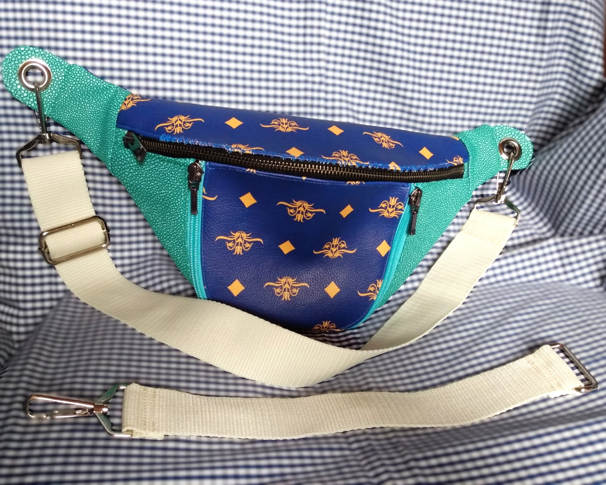 Vue avant du sac banane Mango version bleue. L'extérieur est bicolore en simili cuir émeraude à grains et bleu roi à motifs fleurs et losanges jaunes. Une grande poche principale se ferme via un zip métal. Une poche avant s'ouvre via deux zips bleu clair. La sangle est beige et réglable. Elle s'attache avec des œillets et mousquetons en métal nickelé. Une rallonge de sangle est posée devant la banane. Cette rallonge est beige et contient une boucle d'un côté et un mousqueton de l'autre.