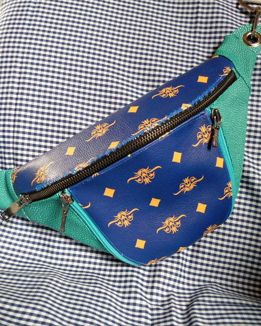 Vue avant du sac banane Mango version bleue. L'extérieur est bicolore en simili cuir émeraude à grains et bleu roi à motifs fleurs et losanges jaunes. Une grande poche principale se ferme via un zip métal. Une poche avant se ferme via deux zips bleu clair. La sangle s'attache avec des œillets et mousquetons en métal nickelé.