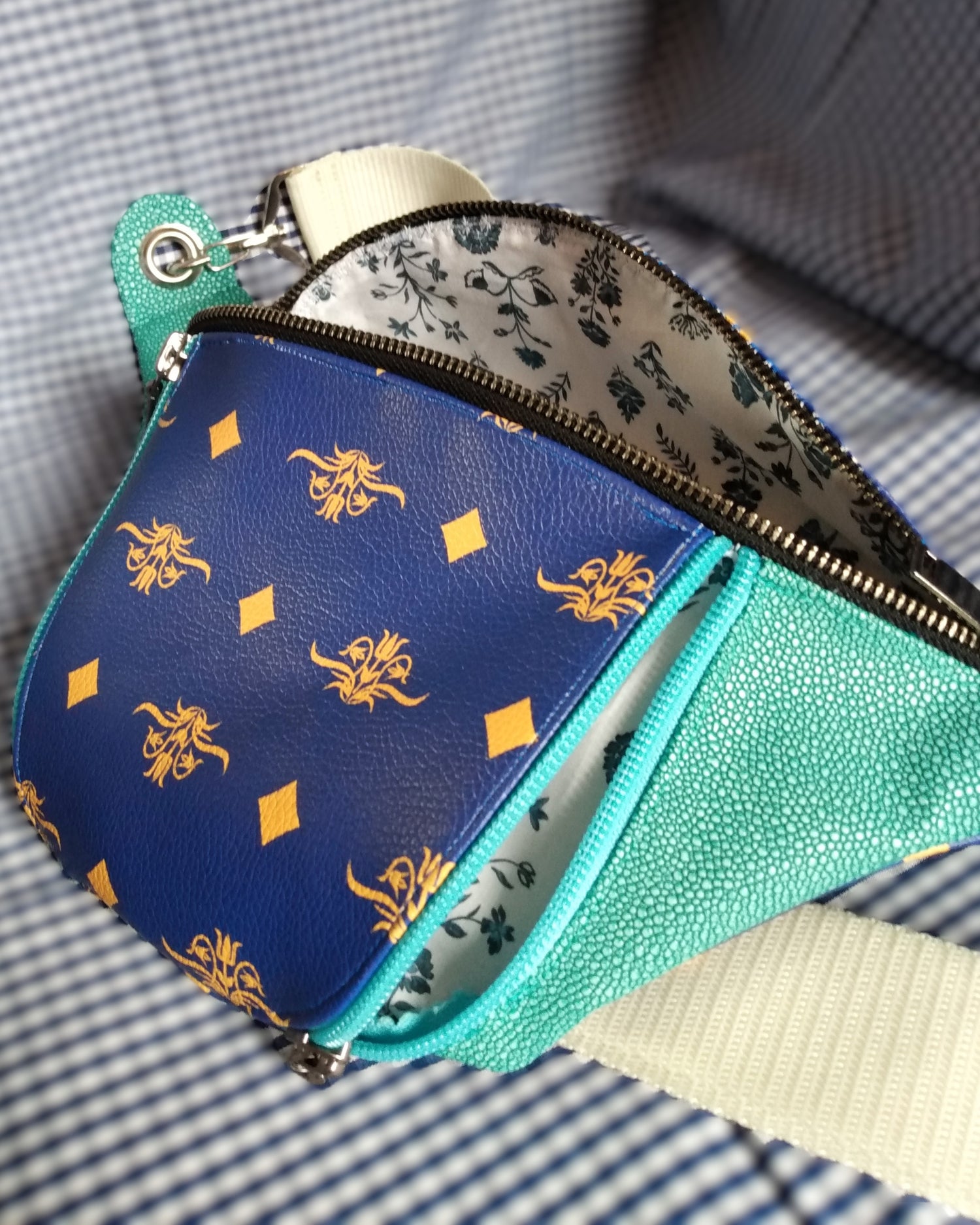 Vue de côté de la banane Mango version bleue. Vue avant du sac banane Mango version bleue. L'extérieur est bicolore en simili cuir émeraude à grains et bleu roi à motifs fleurs et losanges jaunes. Une grande poche principale est ouverte via un zip métal. La poche avant est ouverte par un zip bleu clair. La doublure est blanche à fleurs bleues. La sangle est beige et réglable. Elle s'attache avec des œillets et mousquetons en métal nickelé.