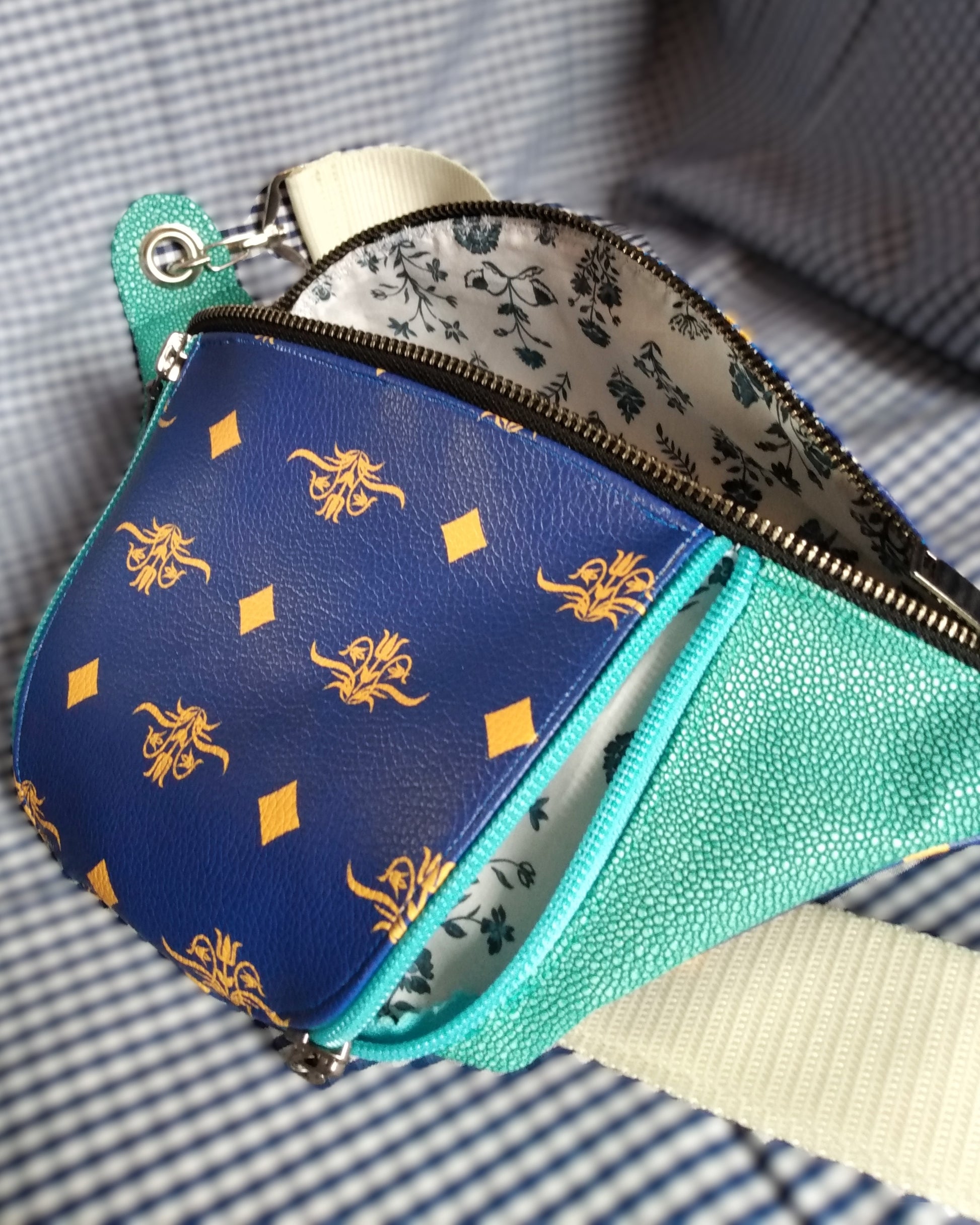 Vue de côté de la banane Mango version bleue. Vue avant du sac banane Mango version bleue. L'extérieur est bicolore en simili cuir émeraude à grains et bleu roi à motifs fleurs et losanges jaunes. Une grande poche principale est ouverte via un zip métal. La poche avant est ouverte par un zip bleu clair. La doublure est blanche à fleurs bleues. La sangle est beige et réglable. Elle s'attache avec des œillets et mousquetons en métal nickelé.