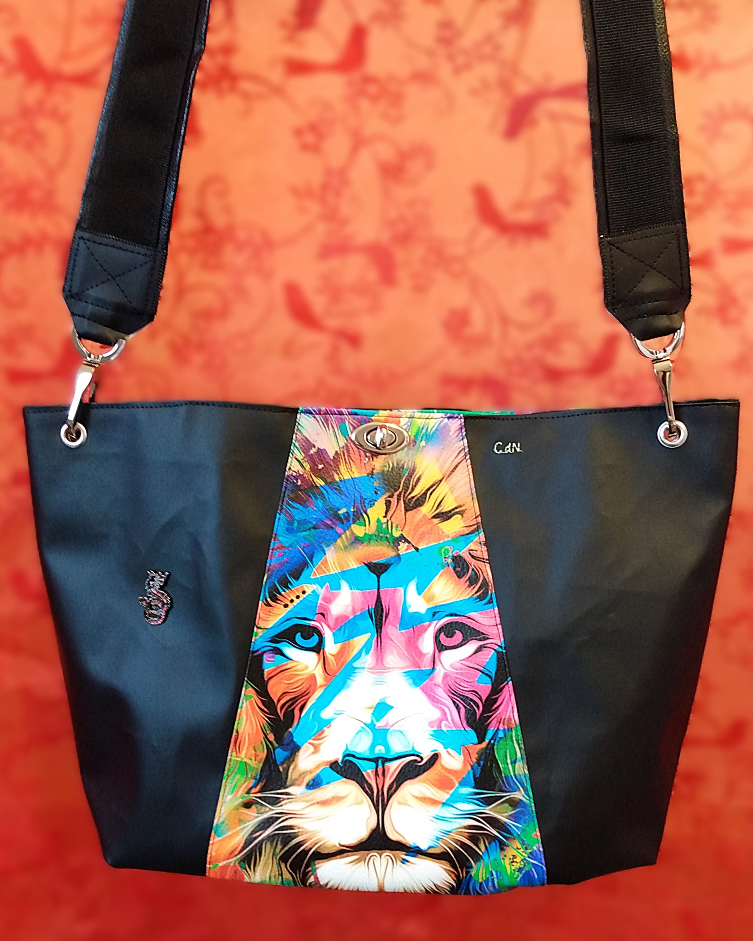 Vue avant du sac Capri en simili cuir. Au centre, une tête de lion multicolore. à gauche et à droite simili cuir noir. A gauche, un pin's en forme de dragon. L'anse est en simili cuir noir et est attachée au sac via des œillets et des mousquetons nickel. Le sac se ferme avec un fermoir tourniquet nickel au centre haut du sac.