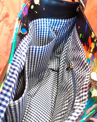Vue intérieure du sac Capri. Doublure en coton à petits carreaux bleus et blancs. On voit, côté arrière, une poche zippée dont la fermeture est bleue clair. Côté arrière, on aperçoit deux poches plaquées.