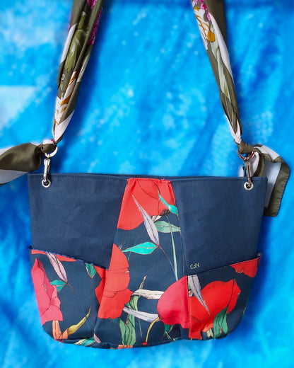 Vue arrière Sac capri fleuri. Le corps du sac est composé de 3 bandes verticales. Au centre une bande en tissu déperlant avec imprimé gros coquelicots rouges et feuilles vertes sur fond bleu foncé. Les 2 bandes latérales sont en tissu épais imperméabilisé bleu foncé. 2 poches latérales sont dans le même tissu qu'au centre. L'anse est un foulard vert kaki et blanc à fleurs roses et blanches. Elle est liée au sac via des mousquetons et œillets nickelés.