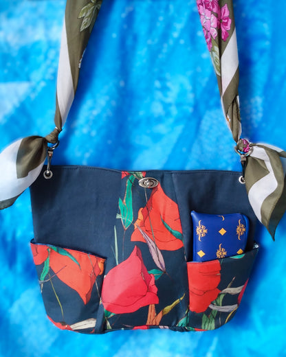 Vue avant Sac capri fleuri. Le corps du sac est composé de 3 bandes verticales. Au centre une bande en tissu déperlant avec imprimé gros coquelicots rouges et feuilles vertes sur fond bleu foncé. Les 2 bandes latérales sont en tissu épais imperméabilisé bleu foncé. 2 poches latérales sont dans le même tissu qu'au centre. Un fermoir tourniquet nickelé ferme le sac. L'anse est un foulard vert kaki et blanc à fleurs roses et blanches. Elle est liée au sac via des mousquetons et œillets nickelés.