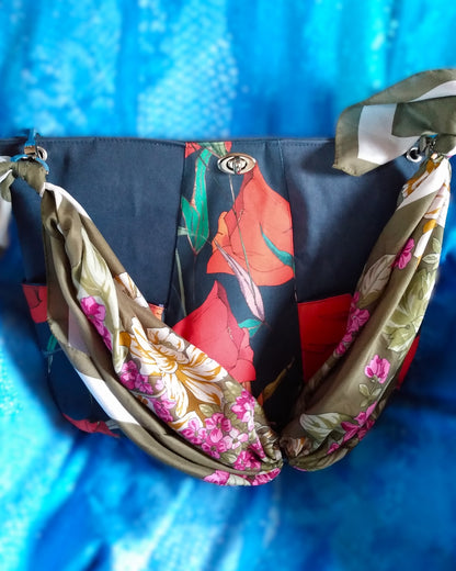 Sac capri fleuri. Le corps du sac est composé de 3 bandes verticales, au centre une bande en tissu déperlant avec imprimé gros coquelicots rouges et feuilles vertes sur fond bleu foncé. Les 2 bandes latérales sont en tissu épais imperméabilisé bleu foncé. 2 poches latérales sont dans le même tissu qu'au centre. Un fermoir tourniquet nickelé ferme le sac. L'anse est un foulard vert kaki et blanc à fleurs roses et blanches. Elle est liée au sac via des mousquetons et œillets nickelés.