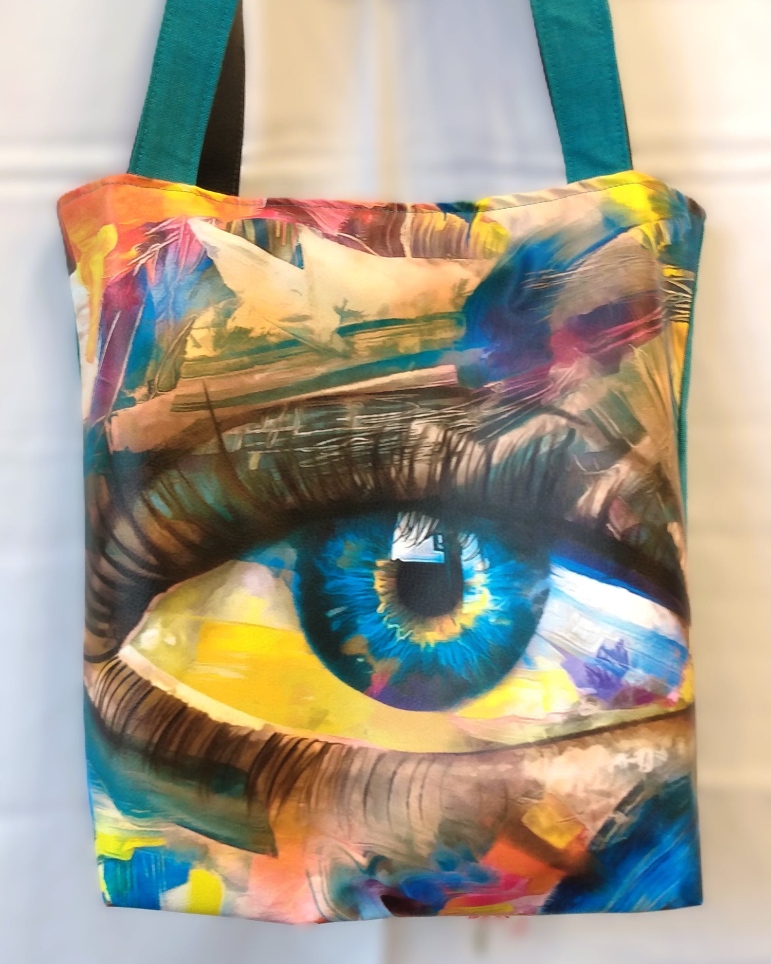 Avant du sac Iris. un gros œil bleu au milieu du sac parait peint. Le haut et bas du sac est multicolore. Les anses sont d'un côté bleues canard et de l'autre grises.