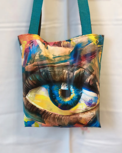 Avant du sac Iris. un gros œil bleu au milieu du sac parait peint. Le haut et bas du sac est multicolore. Les anses sont d'un côté bleues canard et de l'autre grises.