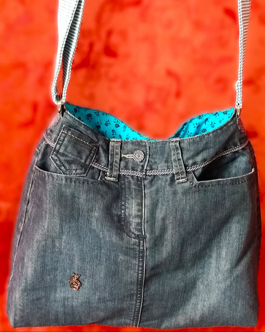 Avant du sac Bleuet. L'extérieur correspond au haut d'un jean, avec deux poches latérales et un bouton rayé blanc et bleu en haut au centre, au-dessus de la braguette. Le jean est bleu/gris. Un pin's en forme de petit dragon est présent vers le bas du sac. La doublure est en coton bleu ciel à petites roses noires. La sangle réglable est en jean rayé bleu et blanc.