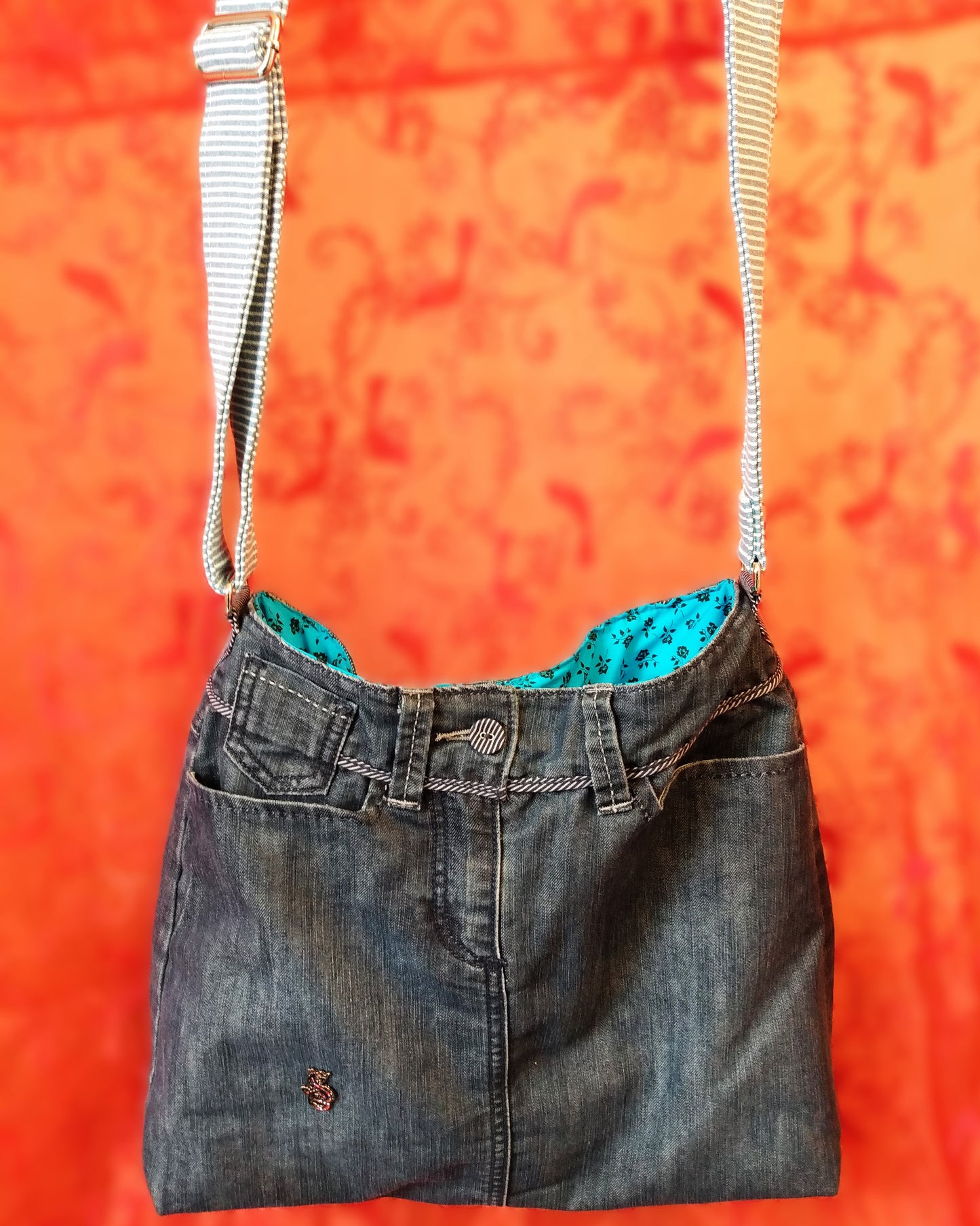 Avant du sac Bleuet. L'extérieur correspond au haut d'un jean, avec deux poches latérales et un bouton rayé blanc et bleu en haut au centre, au-dessus de la braguette. Le jean est bleu/gris. Un pin's en forme de petit dragon est présent vers le bas du sac. La doublure est en coton bleu ciel à petites roses noires. La sangle réglable est en jean rayé bleu et blanc.