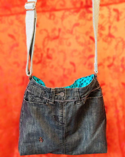 Avant du sac Bleuet. L'extérieur correspond au haut d'un jean, avec deux poches latérales et un bouton rayé blanc et bleu en haut au centre, au-dessus de la braguette. Le jean est bleu/gris. Un pin's en forme de petit dragon est présent vers le bas du sac. La doublure est en coton bleu ciel à petites roses noires. La sangle réglable est en jean rayé bleu et blanc.