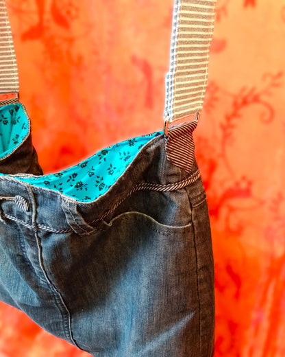 Vue latérale du sac Bleuet. L'extérieur correspond au haut d'un jean. Le jean est bleu/gris. La doublure est en coton bleu ciel à petites roses noires. La sangle réglable en jean à rayures blanches et bleues, est attachée au sac via un anneau rectangulaire.