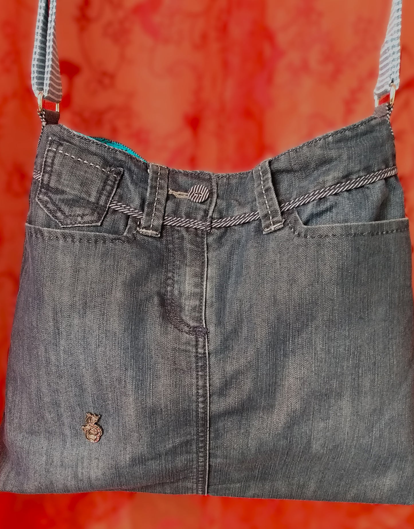 Avant du sac Bleuet. L'extérieur correspond au haut d'un jean, avec deux poches latérales et un bouton rayé blanc et bleu en haut au centre, au-dessus de la braguette. Le jean est bleu/gris. Un pin's en forme de petit dragon est présent vers le bas du sac. La doublure est en coton bleu ciel à petites roses noires. La sangle réglable est en jean rayé bleu et blanc.