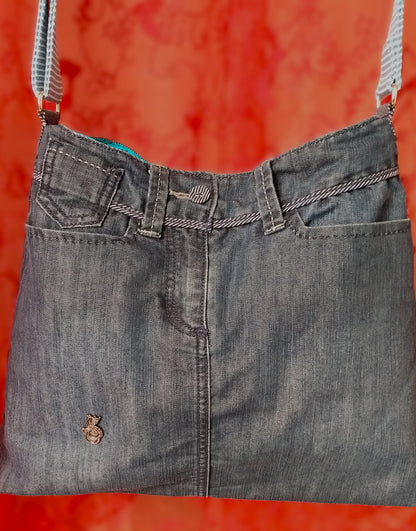 Avant du sac Bleuet. L'extérieur correspond au haut d'un jean, avec deux poches latérales et un bouton rayé blanc et bleu en haut au centre, au-dessus de la braguette. Le jean est bleu/gris. Un pin's en forme de petit dragon est présent vers le bas du sac. La doublure est en coton bleu ciel à petites roses noires. La sangle réglable est en jean rayé bleu et blanc.