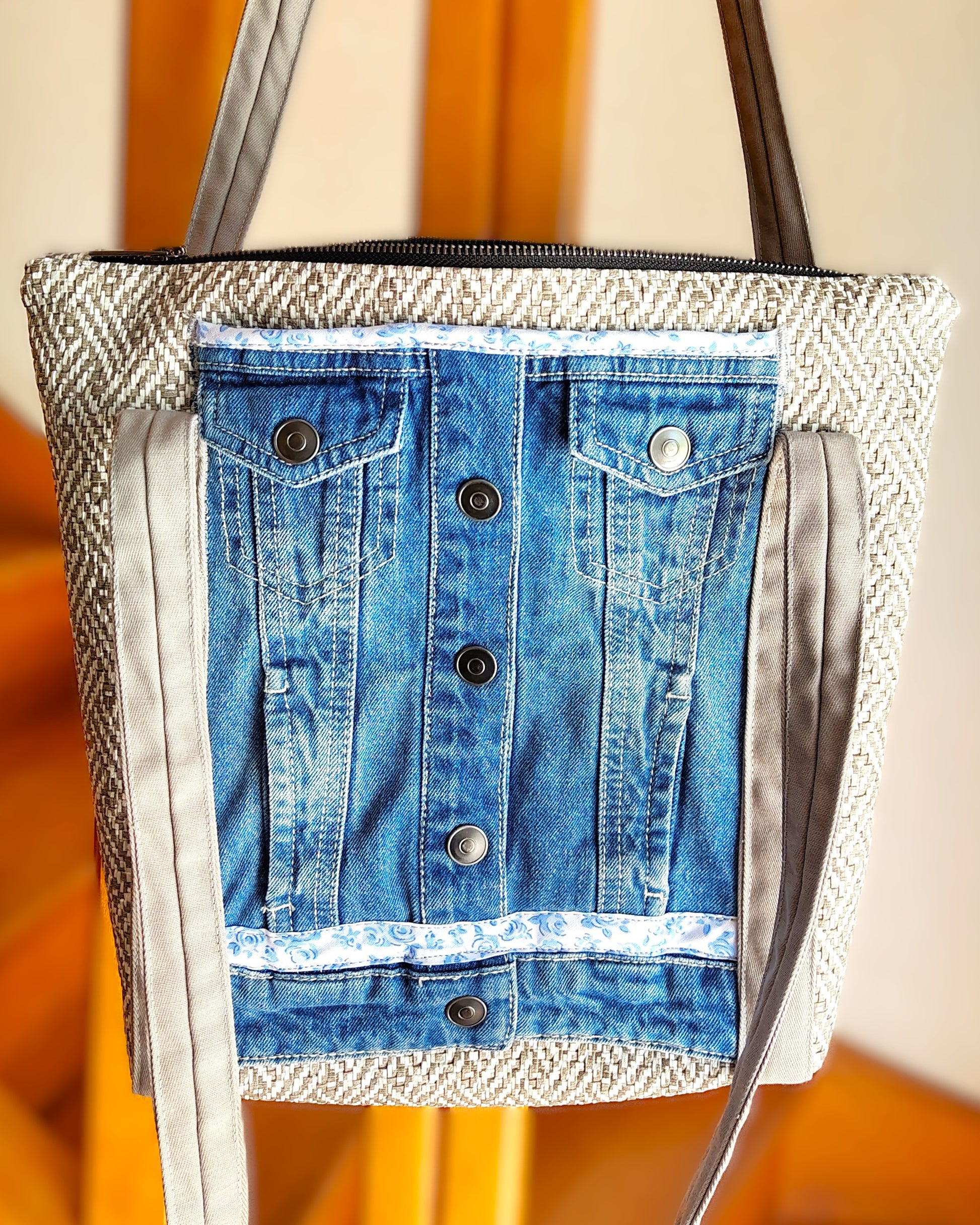 Avant du sac OriJean. tissu principal en simili cuir motif tissage gris et blanc. Grande poche avant fabriquée à partir de l'avant d'une veste en jean bleue. Les anses sont en coton gris clair. Le sac se ferme avec une fermeture éclair métal.