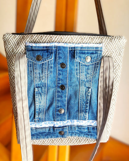 Avant du sac OriJean. tissu principal en simili cuir motif tissage gris et blanc. Grande poche avant fabriquée à partir de l'avant d'une veste en jean bleue. Les anses sont en coton gris clair. Le sac se ferme avec une fermeture éclair métal.