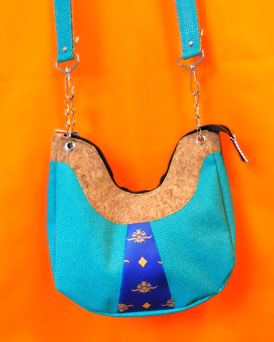 Avant du sac Melbourne. Haut du sac en liège marron clair, sur une bande de environ 8 cm de large. Dessous, le sac a en son centre, un empiècement triangulaire bleu roi à motifs jaunes fleur et losange. Le reste du corps du sac est en simili cuir émeraude à grains. La bandoulière est accrochée via chainettes et mousquetons en métal. Elle est bicolore émeraude à grain et liège. Une fermeture noire permet de fermer le sac.