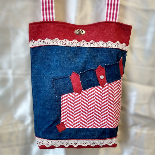 Avant du sac Boujou. Extérieur en denim avec une poche se fermant par un bouton. Fermeture du sac avec un fermoir tourniquet en haut au centre. Anses à rayures rouge et blanche. Bande horizontales en jean rouge en haut (8 cm) et en bas du sac (2 cm). Centre du sac en jean bleu avec une ligne horizontale de dentelle (2cm) en haut et une en bas.
Poche en tissu déperlant à chevrons rouge et blanc, côté gauche du sac. Haut de la poche, alternance de carrés bleus et losanges rouges en jean.