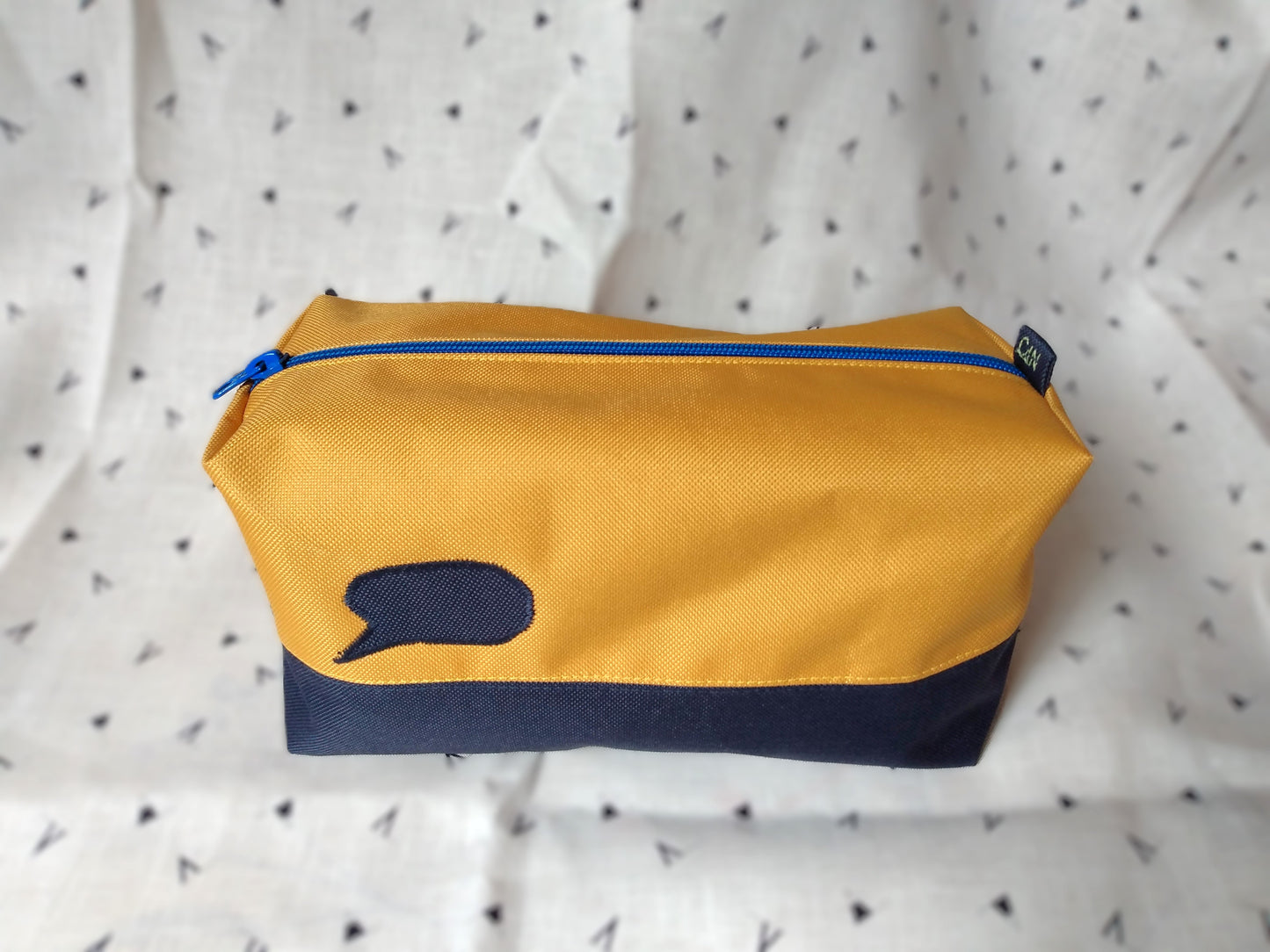 Trousse de toilette Corail rectangulaire et bicolore, en horizontal. bleu foncé en bas et jaune moutarde en haut. Un appliqué bleu foncé en forme de bulle de bande dessinée est présent sur la rayure jaune. Elle se ferme avec une fermeture éclair bleue.