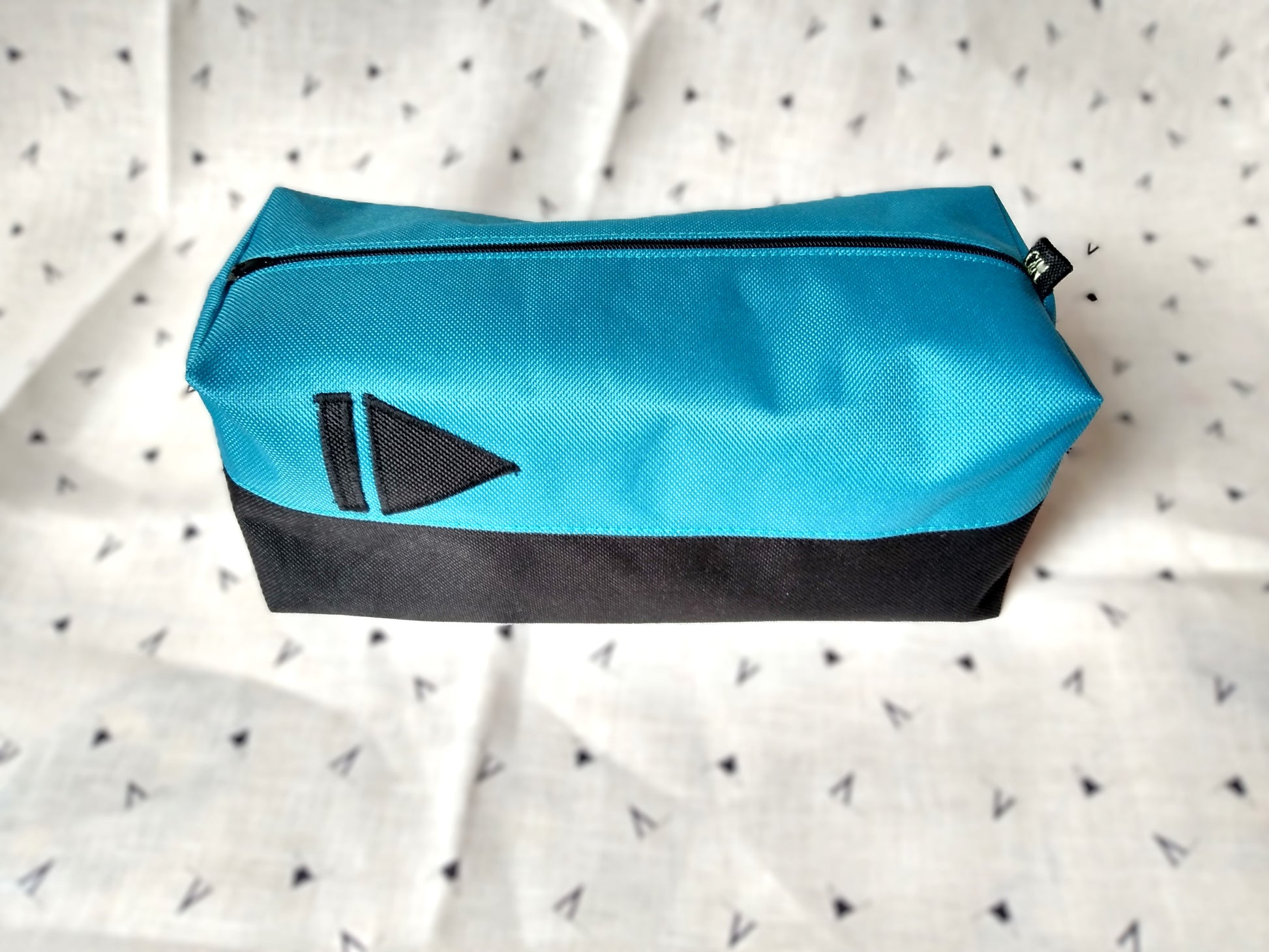 Trousse de toilette Corail rectangulaire et bicolore, en horizontal. Noir en bas et bleu paon en haut. Un appliqué noir en forme de symbole "suivant" (1 trait vertical suivi d'un triangle pointant vers la droite) est présent sur la rayure bleue. Elle se ferme avec une fermeture éclair noire.
