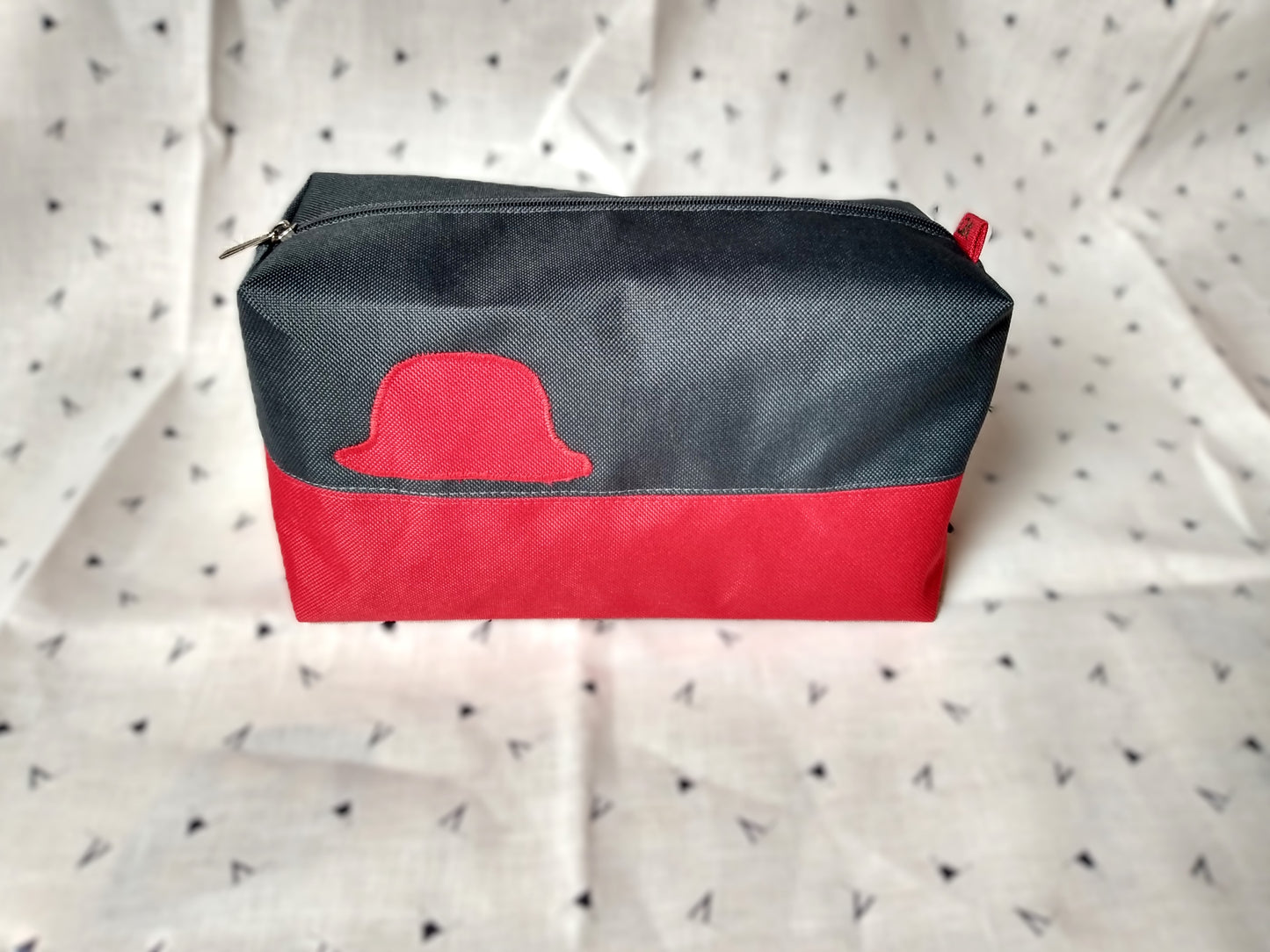 Trousse de toilette Corail rectangulaire et bicolore, en horizontal. Rouge en bas et gris foncé en haut. Un appliqué rouge en forme de chapeau melon est présent sur la rayure grise. Elle se ferme avec une fermeture éclair grise.