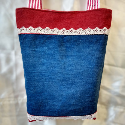 Dos du sac Boujou. Extérieur en denim. Anses à rayures rouge et blanche. Bande horizontales en jean rouge en haut (8 cm) et en bas du sac (2 cm). Centre du sac en jean bleu avec une ligne horizontale de dentelle (2cm) en haut et une en bas.