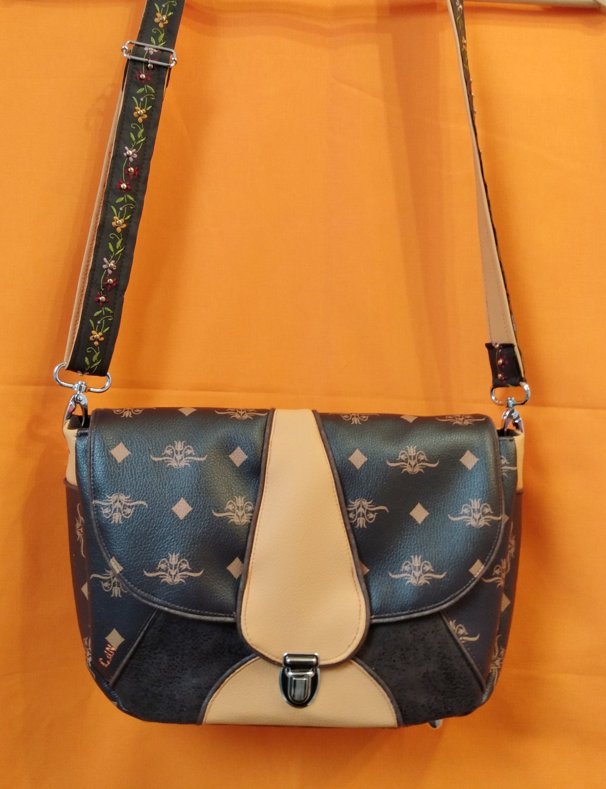 Avant du sac Vegas marron foncé. Extérieur en similicuir marron à fleurs et losanges beiges, abricot et suédine marron. Le rabat, marron et abricot au centre avec passepoil marron, se ferme avec un fermoir cartable. La bandoulière réglable, attachée par deux mousquetons, est en simili orange et coton à petites fleurs brodées. Le corps du sac est marron avec un triangle en suédine marron au centre et un demi cercle orange où est attachée la base du fermoir.