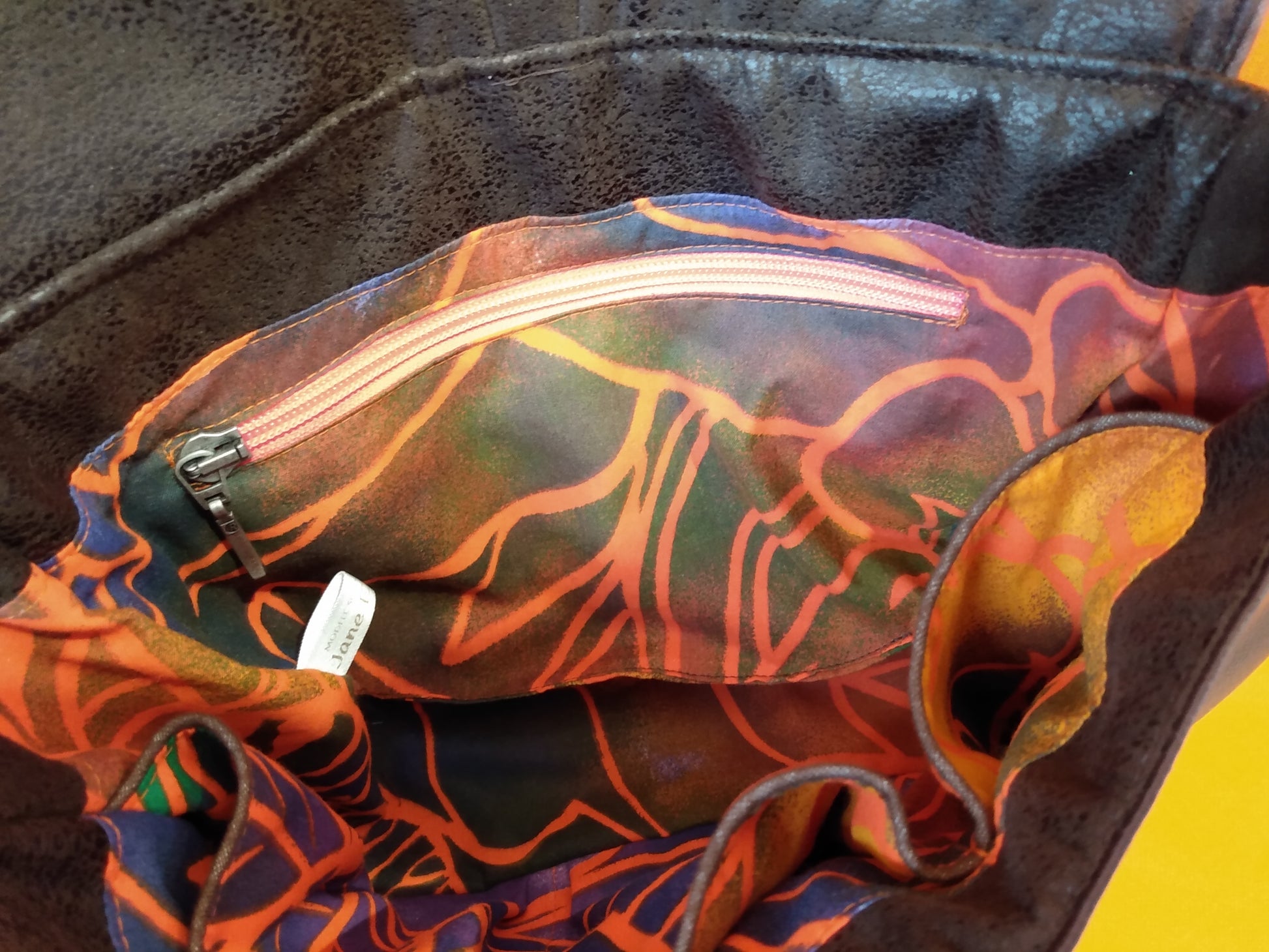 Vue intérieure du sac Vegas marron. coton multicolore à motifs grandes feuilles oranges. Une poche zippée est visible côté dos. La fermeture éclair est rose orangée. côté avant des poches passepoilées sont visibles. Le passepoil est marron.