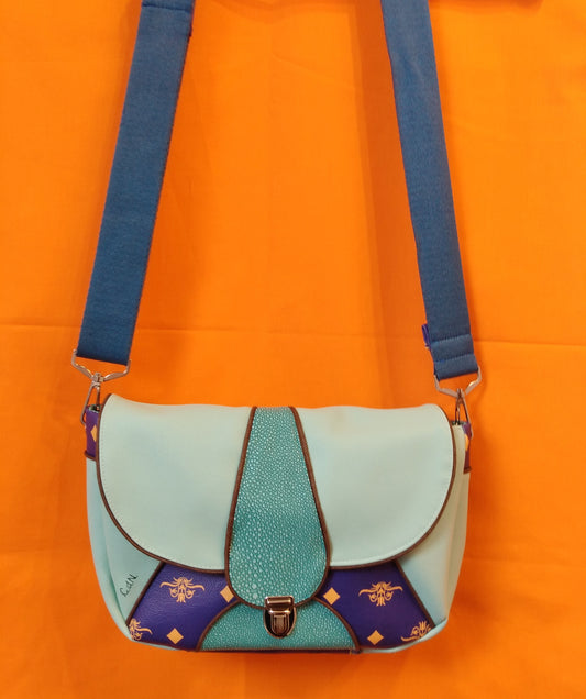 Avant du sac Vegas vert d'eau. Extérieur en similicuir vert d'eau uni, émeraude à grains et bleu roi à motifs fleurs et losanges jaunes. Le rabat, vert d'eau et émeraude au centre avec passepoil marron, se ferme avec un fermoir cartable. La bandoulière réglable, attachée par deux mousquetons, est en coton bleu. Le corps du sac est vert d'eau avec un triangle bleu roi au centre et un demi cercle émeraude où est attachée la base du fermoir.